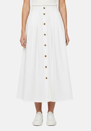 Donna che indossa una gonna midi bianca con bottoni sul davanti abbinata a sneaker bianche, in piedi davanti a uno sfondo semplice.