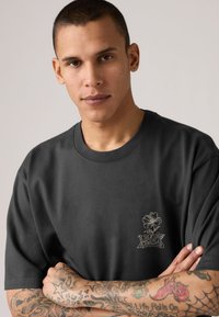 Νέος με κοντά μαλλιά που φοράει σκούρο γκρι μπλουζάκι με λουλουδάτο σχέδιο και κείμενο, με τα χέρια σταυρωμένα, εμφανίζοντας πολύχρωμα τατουάζ στα κάτω μπράτσα.