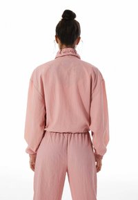 Světle růžová, oversized bunda s volánovým límcem a elastickými manžetami, spárovaná s kalhotami na gumě z odpovídající látky, která má strukturovaný povrch.