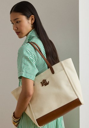 Kvinna med långt mörkt hår, iklädd grönrandig skjorta och guldfärgade armband, som håller en stor beige tygväska med bruna läderdetaljer.