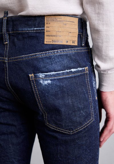 Nahaufnahme einer Person, die dunkelblaue Jeans trägt. Die hintere Tasche ist abgewetzt und darüber befindet sich ein braunes Lederetikett.