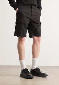 Svart cargoshorts med frontlommer, kombinert med hvite sokker og svarte snørestøvler. Stoffet ser slitesterkt og glatt ut.