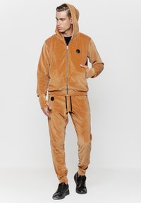 Beżowy dres z weluru, w skład którego wchodzi rozpinana bluza z kapturem oraz dresowe spodnie. Posiada okrągły, czarny detal z logo. Materiał ma miękką, puszystą teksturę.