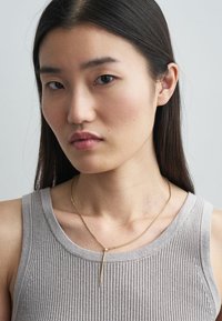 Colar dourado com corrente fina e pendente de barra vertical, usado sobre um top cinza claro canelado. Textura lisa, design minimalista.