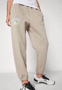 Helle beige Jogginghosen mit elastischen Bündchen, die über ein grafisches Logo an den Oberschenkeln und eine Reißverschlusstasche mit limegrünem Akzent verfügen.