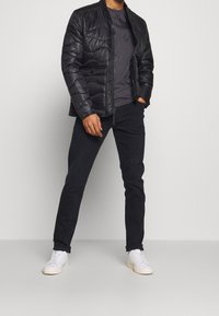 Giacca padded nera con zip, indossata sopra una camicia a fantasia scura, abbinata a pantaloni scuri e sneakers bianche. Design pulito e lineare.