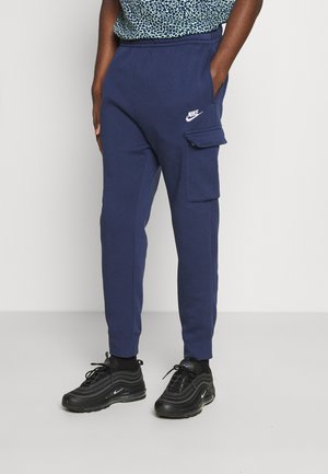 Osoba ubrana w granatowe spodnie dresowe Nike z kieszenią typu cargo i czarne sneakersy Nike, stojąca na jednolitym jasnym tle.