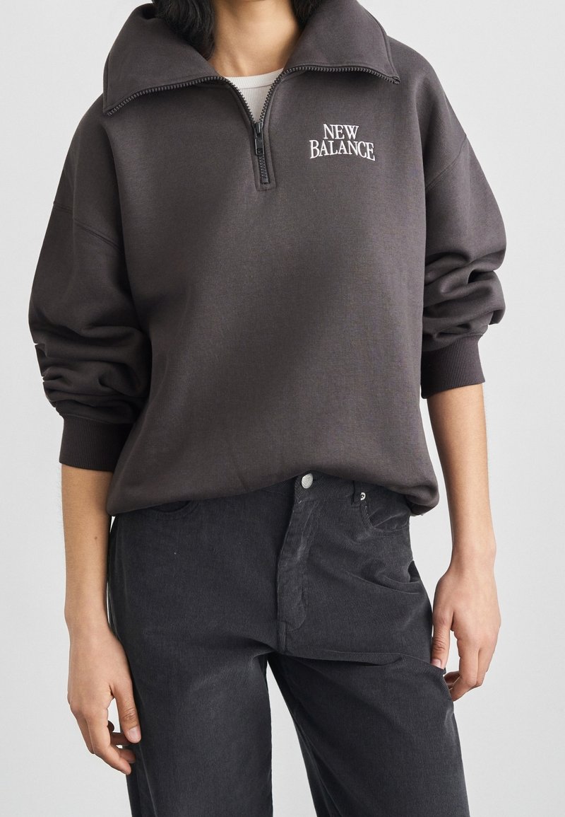 Personne portant un sweat-shirt New Balance gris foncé à fermeture éclair quart de zip, manches retroussées, et un pantalon noir sur un fond uni.