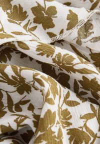 Tissu à motifs floraux en crème et vert olive, présentant un tissage doux et texturé avec des bords effilochés et des feuilles et fleurs entrelacées.