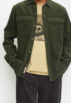 Mann trägt ein dunkles, grünes Cordhemd über einem beigefarbenen T-Shirt mit Grafik und dunkle Cordhosen, Hände in den Taschen.