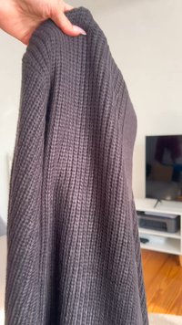 Tumši pelēks adīts džemperis, ko tur rokās, ar teksturētu, ribota raksta dizainu. Tam ir brīvs piegriezums un pagarināta silueta forma.