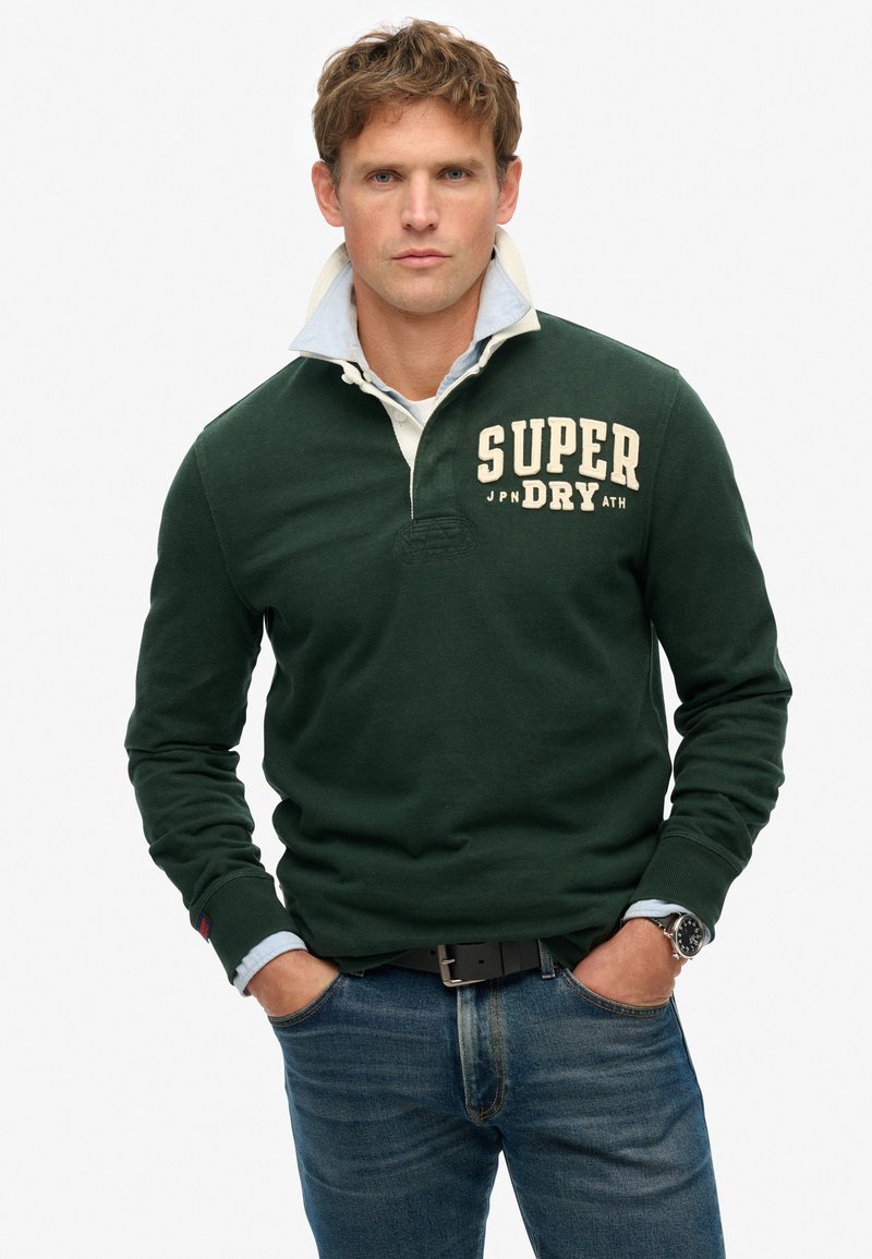 Donkergroen polo met lange mouwen en een lichtblauw overhemd eronder, met een crèmekleurig logo. Gecombineerd met denim jeans en een zwarte riem.
