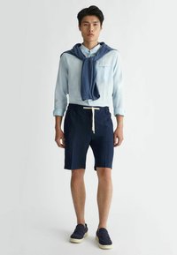 Camicia azzurra chiara con bottoni, pantaloni corti blu navy con cordoncino, maglione blu scuro legato attorno al collo e scarpe nere slip-on.