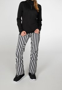 Person i svart långärmad tröja och svart-vita vertikalt randiga byxor, iförd svarta sneakers med vita sulor, står mot en enfärgad bakgrund.