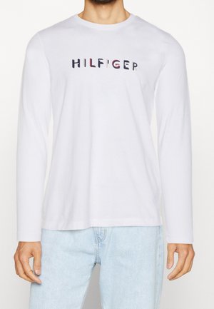 Homme portant une chemise blanche à manches longues avec un texte coloré "HILFIGER" sur la poitrine et un jean bleu clair, visible du cou aux hanches.