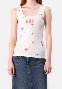 Débardeur blanc à côtes avec un col rond, orné de motifs floraux multicolores et du logo "GAP" en rose, porté avec une jupe en denim.