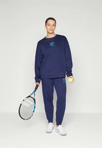 Teget plava majica s kapuljačom i odgovarajuće hlače od mekanog tkanja, s logotipom u tirkiznoj boji. Pribor uključuje tenisku lopticu i reket.