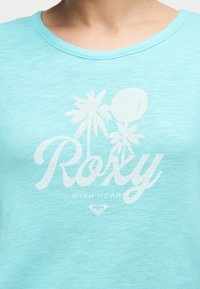 Ανοιχτό μπλε πουκάμισο με λευκά φοίνικες, τη λέξη «Roxy» και τη φράση «With Heart» τυπωμένα μπροστά.