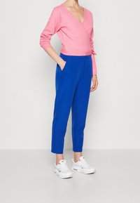 Light pink wrap sweater with long sleeves paired with blue tapered pants. Model wears white sneakers, showcasing a contrast in colors and textures.