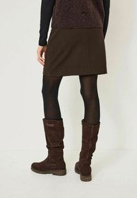 Bottes en suède marron jusqu'au genou avec une pointe arrondie, un tissu texturé et une semelle en caoutchouc ; portées avec une jupe courte marron et des collants noirs.