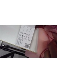 Étiquette d'entretien des vêtements avec symboles de lavage et informations sur la taille tenue à la main, attachée à un tissu rose sur une surface blanche.