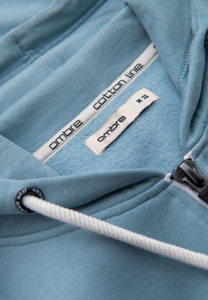 Sudadera de algodón de color azul claro con un suave forro interior, cierre con cremallera y cordones blancos con detalles en negro. Etiquetas visibles en el interior.