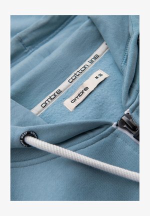 Světle modrá bavlněná mikina s měkkou vnitřní podšívkou, zipovým zapínáním a bílými šňůrkami s černými akcenty. Vnitřní štítky jsou viditelné.