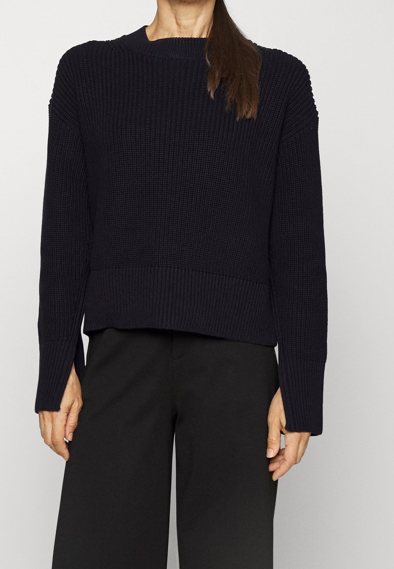 Pull en tricot bleu marine avec col rond, motifs texturés, coupe courte et poignets côtelés. Associé à un pantalon noir à jambes larges.