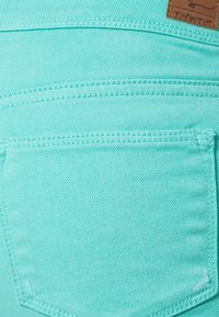 Akvapain denimkangas, jossa on sileä rakenne, takataskut ja kontrastiväriset ompeleet. Ruskea nahkalappu vyötäröllä.