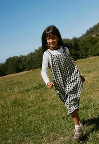 Jeune fille en robe longue à carreaux courant sur l'herbe verte avec des arbres et un ciel bleu clair en arrière-plan.