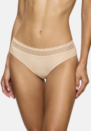 Femme portant un sous-vêtement style bikini beige orné de dentelle, debout avec les bras détendus le long du corps sur un fond uni.