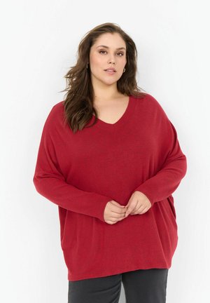 Roter übergroßer Pullover mit V-Ausschnitt, langen Ärmeln und entspanntem Schnitt. Weicher Stoff mit subtiler Textur, kombiniert mit dunklen Hosen.