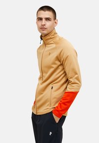 Tan zip-up jakke med orange ærmer, fremstillet af blødt stof. Har en høj krave, sidelommer og et logo på brystet.