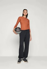 Oransje langermet topp med en halv glidelås, kombinert med svarte vide joggebukser og svarte sneakers. Holder en svart basketball med en rosa logo.