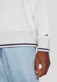 Felpa grigia chiara con polsini a coste, caratterizzata da strisce blu e bianche, abbinata a jeans in denim blu, con un'attenzione particolare ai dettagli delle maniche.