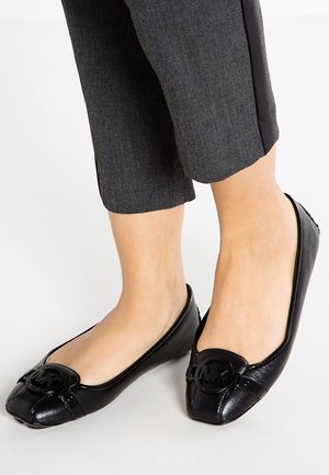 Osoba nosząca czarne, płaskie buty Michael Kors ze skórzaną cholewką i logo oraz przycięte, ciemnoszare spodnie, stojąca na białej powierzchni.