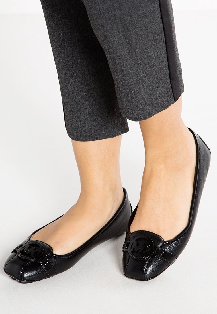 Personne portant des chaussures plates en cuir noir Michael Kors avec emblème du logo et un pantalon gris foncé raccourci, debout sur une surface blanche.