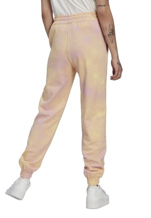 Persona vestida con pantalones jogger teñidos de rosa claro y amarillo con puños elásticos y zapatillas blancas, vista desde atrás.