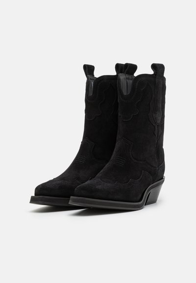 Botas vaqueras de ante negro con puntera puntiaguda, detalles de costura curvada, asas de tiro y un tacón bajo y apilado. Forro de cuero suave.