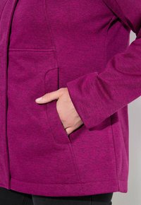 Ręka w bocznej kieszeni fioletowej, teksturowanej kurtki z długim rękawem, z czarnymi spodniami widocznymi na dole.