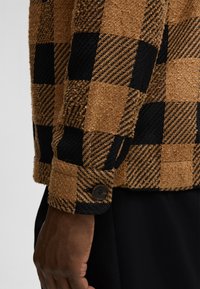Bruine en zwarte geblokte jas van een textuurstof met een fuzzy uitstraling. Kenmerkt een knoopmanchet detail en contrasterende strepen. Warm, gelaagd ontwerp.