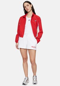 Cazadora roja con acentos en azul y beige, camiseta sin mangas blanca, pantalones cortos blancos con texto rojo "R.S.V.P.", y zapatillas plateadas. Texturas de tela suaves.