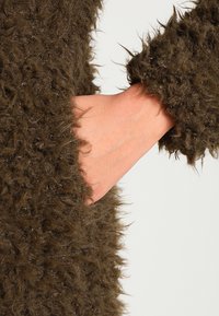 Brązowa, puszysta tkanina o teksturowanym, kudłatym wyglądzie. Jedna ręka częściowo widoczna, odsłaniająca gładką nadgarstek w zestawieniu z płaszczem.