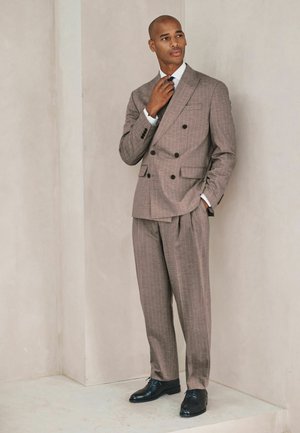 Mann, der seine Krawatte richtet, trägt einen braunen, zweireihigen Anzug mit dezenten Streifen und schwarze Anzugschuhe, steht vor einer hellbeigen Wand.