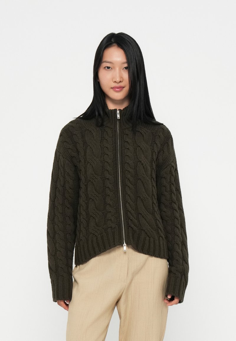Dunkelgrüner Kabelstrickpullover mit hohem Kragen und silbernem Reißverschluss. Gerippter Saum und Bündchen. Trägt eine hellfarbige Hose.