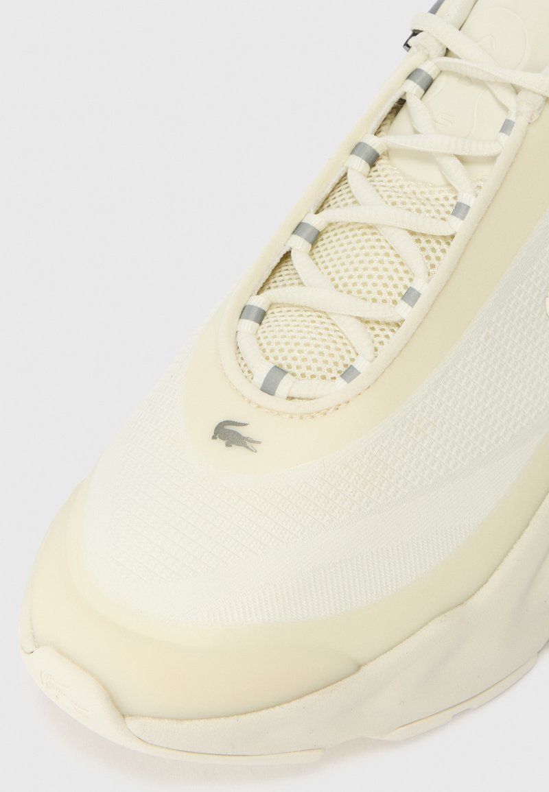 Lacoste Sneaker in Off-White mit einer strukturierten Mesh-Oberfläche, geschichtetem Design und grauen Akzenten an den Schnürsenkeln und dem Logo in der Nähe der Zehen.