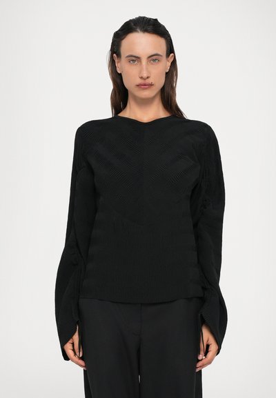 Vrouw met recht, donker haar draagt een gestructureerde zwarte top met lange mouwen en brede manchetten, met bijpassende zwarte broek, en kijkt recht vooruit.