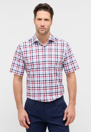 Uomo che indossa una camicia a maniche corte con motivo a quadri rosso, blu e bianco e pantaloni blu scuro, in piedi davanti a uno sfondo semplice.