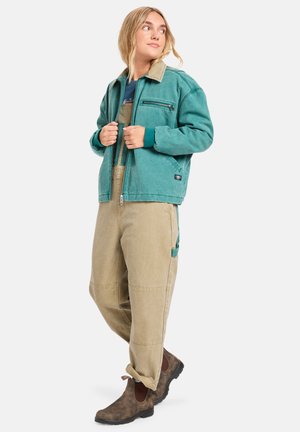 Teal jakke med en beige krage og foran glidelåslommer, kombinert med beige overaller. Brune ankelstøvler fullfører antrekket.