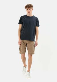 Navy cotton t-shirt paired with tan cargo shorts featuring two side pockets and white sneakers. Model stands on a plain backdrop.